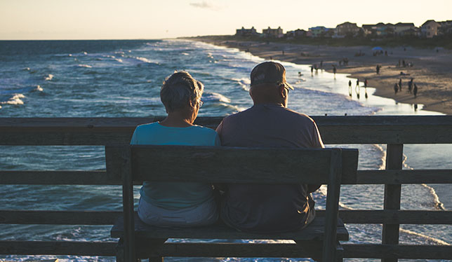 An elderly couple sitting on a bench overlooking the ocean.