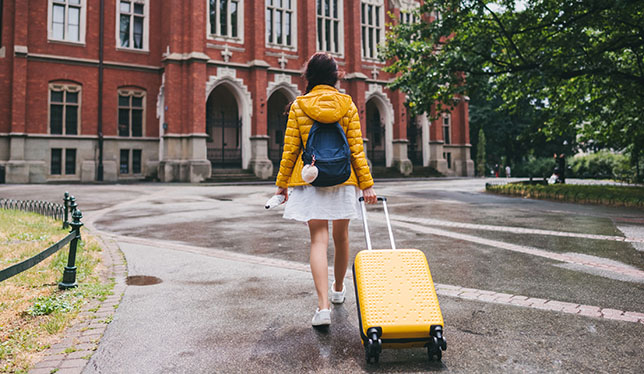 Girl in yellow jacket with suitcase walking towards a university campus.